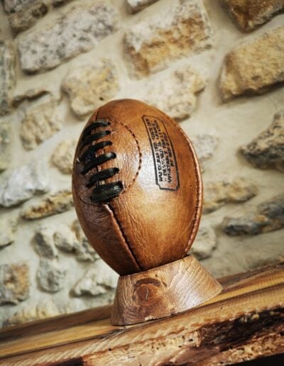 Un mini ballon de rugby posé sur son socle en bois devant un mur en pierres de taille.