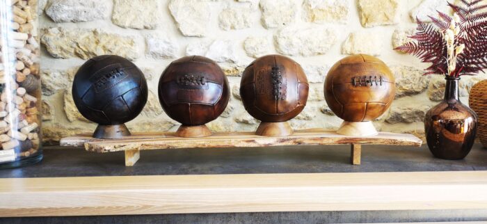 Une gamme de 4 ballons de foot vintage posés sur leur socle en bois le tout présenté sur un meuble en bois.