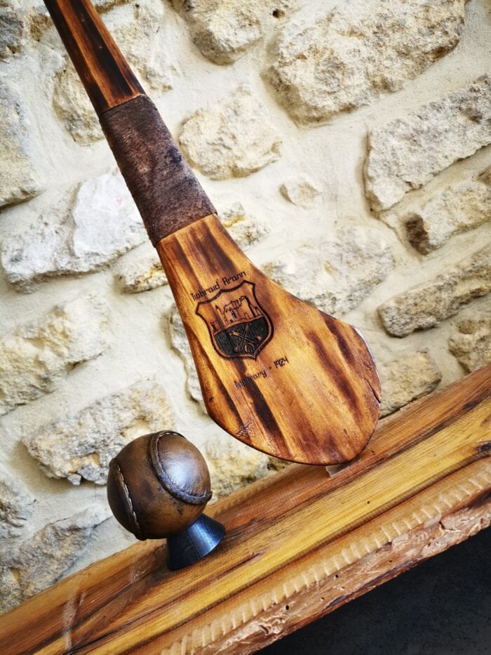 Photo d'une crosse de hurling foncée posée sur un meuble en bois, à côté d'un sliotar